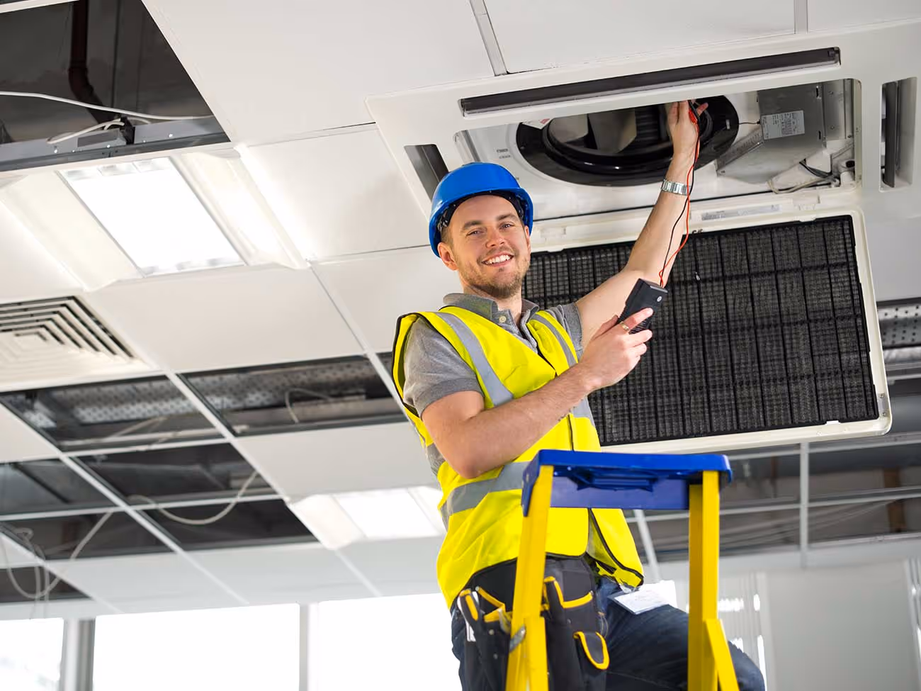 Techniker mit Schutzhelm und Warnweste überprüft eine Klimaanlage an der Decke.